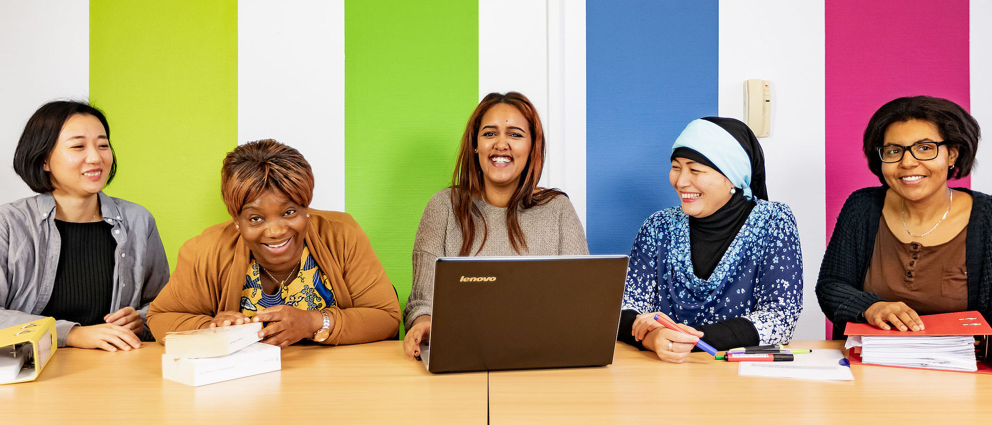 Fünf Frauen sitzen vor einer bunt gestreiften Wand an einem Tisch mit Laptops und Ordnern