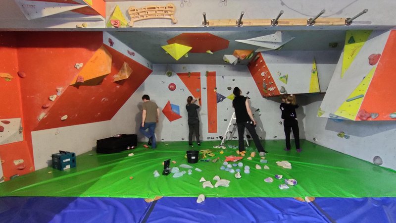 4 Menschen stehen in einer Boulderhalle an der Wand und schrauben Griffe ab. Im Vordergrund liegen viele Griffe auf den Matten.
