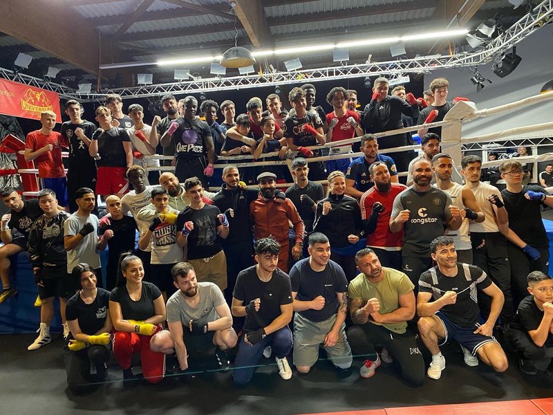 Junge Menschen stehen oder hocken zusammen für das Gruppenfoto vor einem Boxring und heben die Fäuste hoch