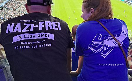 Zwei Menschen von hinten im Stadion, beide tragen T-Shirts der Schalker Fan-Initiative 