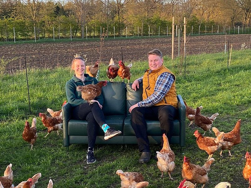 Eine und Frau und ein Mann sitzen auf einem grünen Sofa auf einer Wiese, um sie herum laufen Hühner