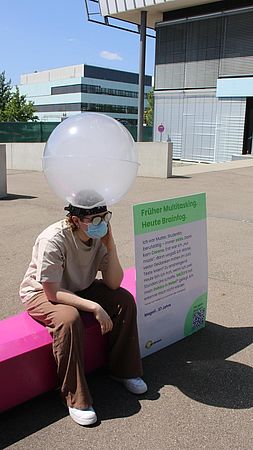 Person trägt eine mit Trockennebel gefüllte Kugel auf dem Kopf. Hinter der Person wird auf einem Plakat das Symptom Brainfog bei ME/CFS erklärt.