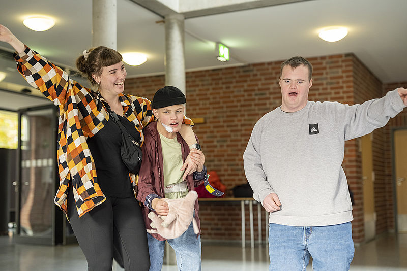 Eine Frau und zwei junge Männer stehen nebeneinander. Die Frau hält einen Jungen im Arm und hebt den anderen Arm lachend hoch. Der andere Junge hält seinen Arm auch hoch.