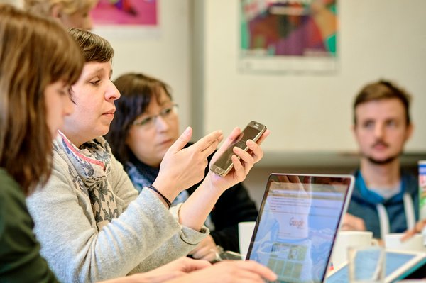 Vier Personen sitzen nebeneinander. Ein aufgeklappter Laptop steht vor ihnen auf dem Tisch. Eine Frau hält ein Smartphone in der Hand. 