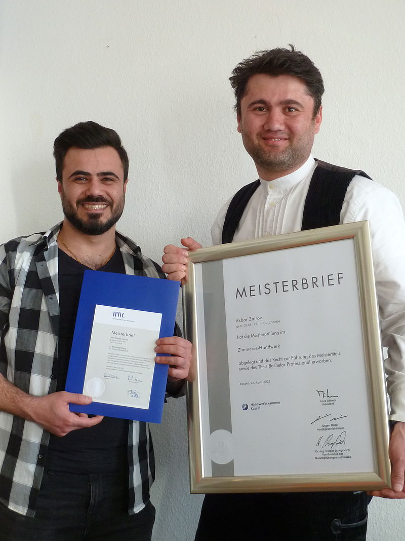 Sherak Wanes (Frisör) aus Syrien und Akbar Zoirov (Zimmerer) aus Tadschikistan stehen nebeneinander und zeigen ihre Meisterbriefe