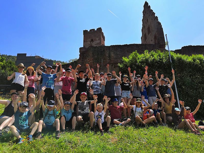Kinder und Jugendliche stehen und sitzen für ein Gruppenfoto vor einer Burg und winken in die Kamera