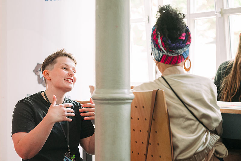 Queermed Gründer*in Sara Grzybek hockt vor einer Teilnehmenden bei einem Event und spricht mit dieser