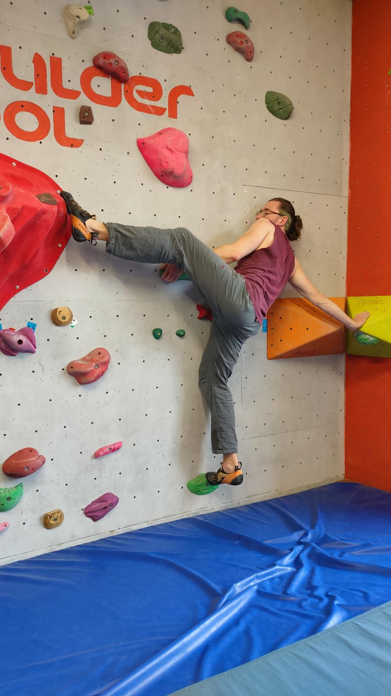 Ein junger Mann klettert an einer Boulderwand. Mit der rechten Hand stützt er sich stark auf ein Volumen, der linke Fuß ist etwa auf Schulterhöhe auf einem Griff.