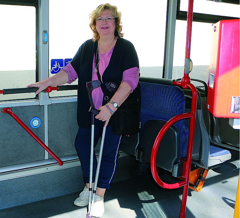 Frau steht im Bus und hält sich mit einer Hand fest. In der anderen Hand hält sie Krücken.