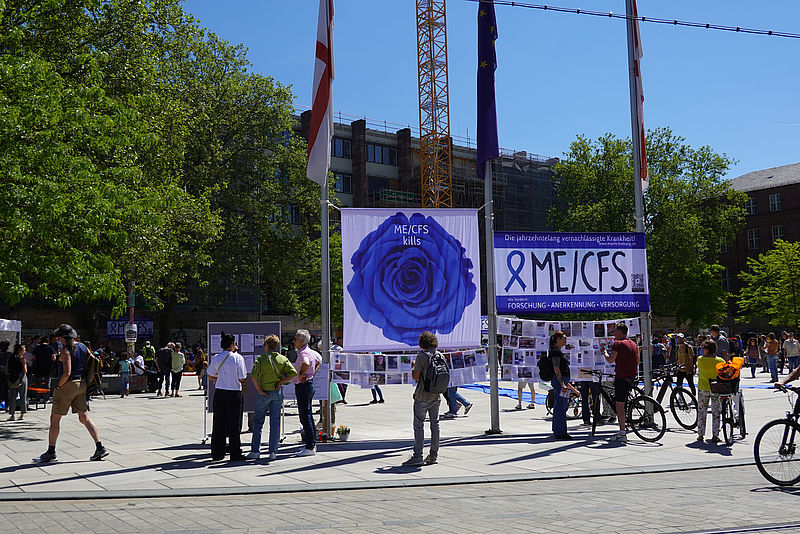 Blick auf den Platz der Alten Synagoge. Zwischen Fahnenmasten sind zwei große Banner und viele kleinere Botschaften von Betroffenen aufgehängt. Passanten lesen diese interessiert. Das eine Banner zeigt eine große blauer Rose mit dem Text "ME/CFS kills". Das andere Banner hat die Aufschrift "ME/CFS - Die jahrzehnte vernachlässigte Krankheit. Wir fordern Forschung, Anerkennung, Versorgung".