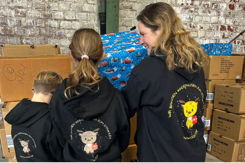 Drei Ehrenamtliche mit GGKT-Hoodies stehen lächelnd vor Kartons und verpackten Geschenken in einem Lager.