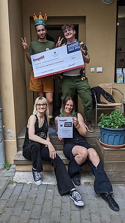 vier Menschen auf der Treppe zu einer Terrasse, die vorderen beiden sitzen auf den Stufen der Treppe, die rechts hält die Urkunde des Jugendengagementpreises, die beiden hinteren stehen auf der Treppe und halten den Scheck für den Jugendengagementpreis, der linke trägt die Regenbogenkrone für den Preis
