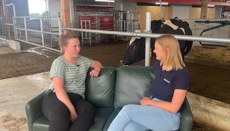 Zwei junge Frauen sitzen auf einem grünen Sofa, das in einem Stall vor Kühen steht.