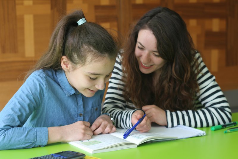 Eine Mentorin sitzt mit ihrer Schülerin am Tisch und hilft ihr beim Lesen eines Buches.