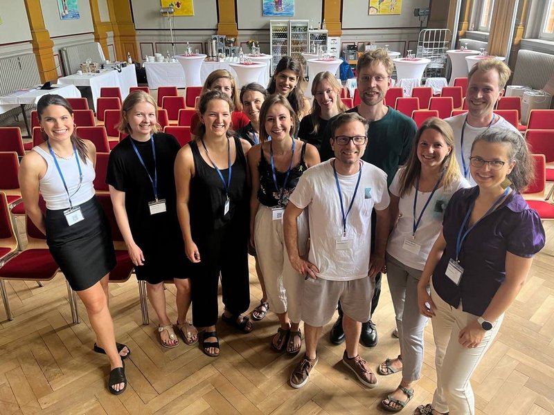 Eine Gruppe junger Menschen steht in einem Raum vor Stühlen und schaut in die Kamera