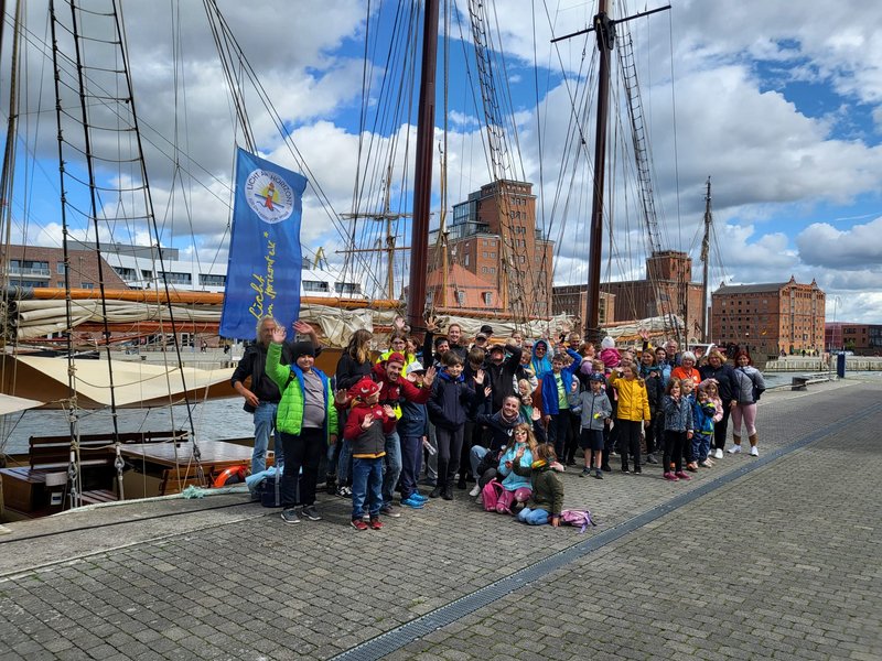 Gruppenfoto von Kindern, Familien und Vereinsmitgliedern am Hafen von Wismar vor einem Segelschiff mit Vereinsfahne.