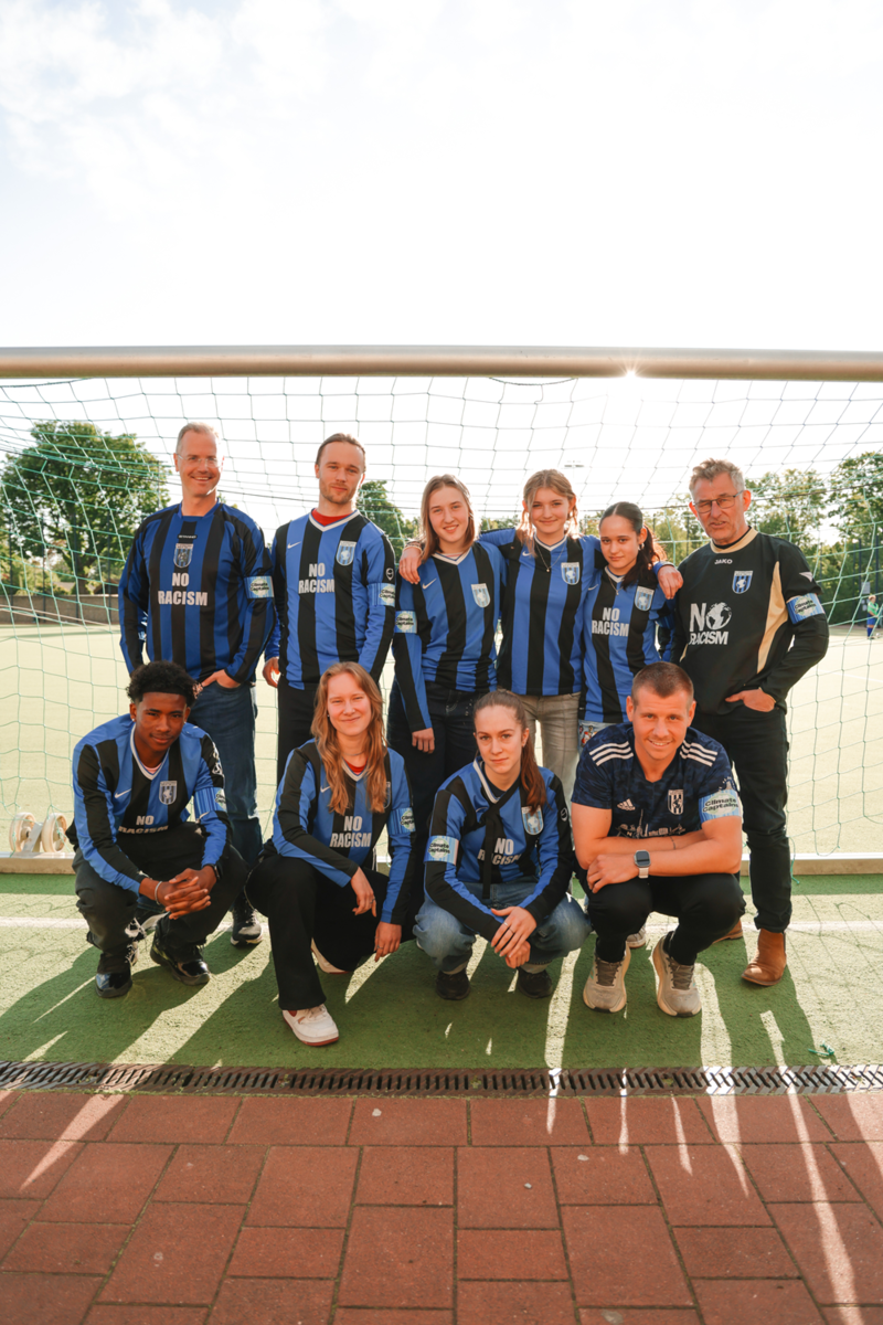 Zehn Personen stehen und hocken in einem Fußballtor und bilden ein Team, dabei tragen sie eine Binde mit den Climate Stripes.