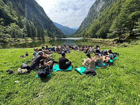 Auf dem Bild sieht man Teilnehmende der Sommerakademie in einer großen Runde auf einer Wiese sitzen, im Hintergrund eine beeindruckende Kulisse aus Bergen und Bergsee.
