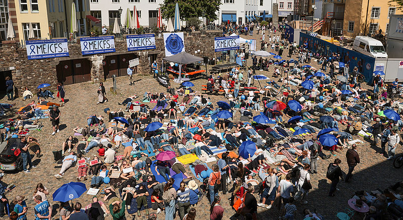 Der Augustinerplatz ist gefüllt mit Menschen, die liegend demonstrieren. Auf dem Platz sind mehreren ME/CFS-Banner aufgehängt (Beschreibung der Bannner siehe Bild 2).
