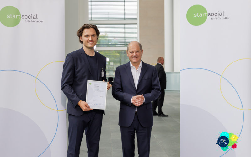 Man sieht zwei Männer nebeneinander stehen. Einer hält stolz eine Urkunde. Der andere Mann ist der Bundeskanzler Olaf Scholz. Er schaut freundlich in die Kamera.