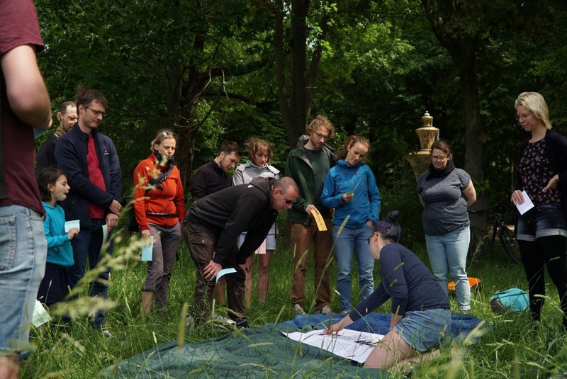 Eine Teamerin deutet auf ein Poster, das auf einer Decke ausgebreitet ist. Einige Menschen stehen rundherum auf einer Wiese und betrachten das Poster interessiert.